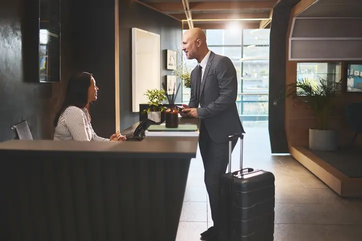 Receptionist, people and man in hotel with check in for accommodation on business trip. Woman, employee and male client or customer with smile in help desk for booking and hospitality as guest